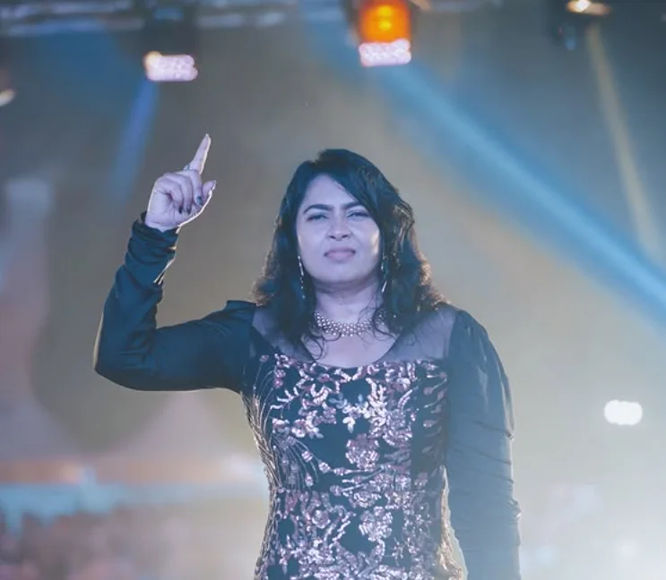 Reena on stage in a sequinned gown, one finger raised to the audience