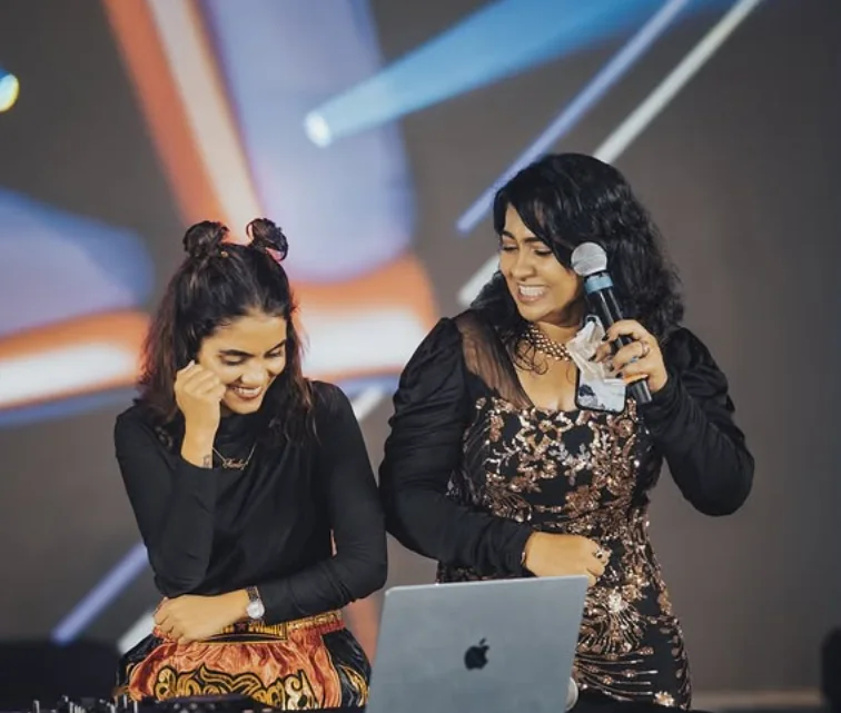 Reena laughing on stage with a microphone beside a DJ at her laptop